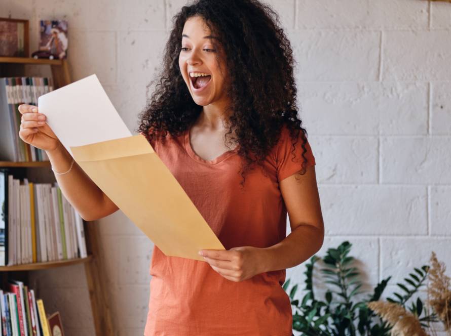 Young joyful woman with dark curly hair in T-shirt happily opening envelope with exam results letter